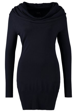 Femme portant une robe noire à manches longues avec col bénitier, des collants noirs, des bottines taupe, tenant un sac à main marron texturé, debout sur un fond clair.