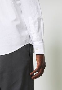 Bras droit d'une personne portant une chemise blanche à manches longues et un pantalon gris foncé, la main détendue le long de la cuisse.