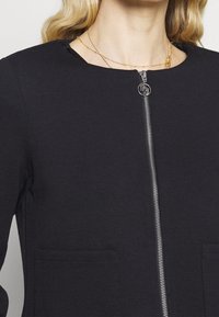 Czarna kurtka zapinana na zamek błyskawiczny z srebrnym zamkiem i dwoma przednimi kieszeniami. Materiał jest gładki, a design posiada zaokrąglony dekolt.