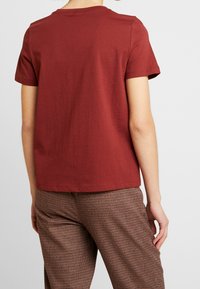 Kortærmet t-shirt med rund hals i rødbrunt bomuldsstof. Har en afslappet pasform, glat tekstur og syninger rundt om halsudskæringen.