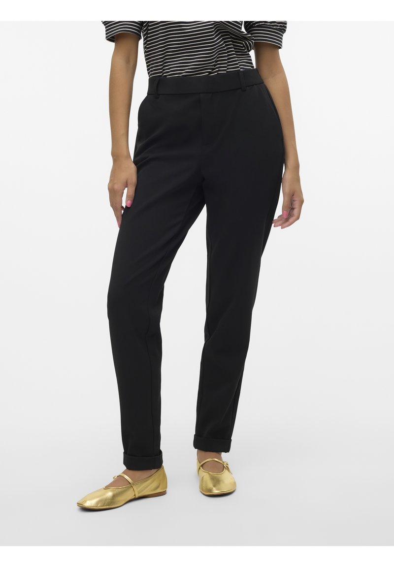 Femme portant un pantalon noir taille haute avec ourlets retroussés, un haut à manches courtes rayé et des ballerines Mary Jane dorées, debout devant un fond blanc.