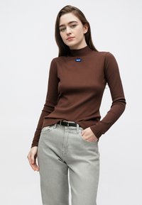 Pruun pika varrukaga soonikkoes pluus kõrge kaelusega ja sinise logomärgiga, kombineeritud helehallide teksade ja musta vöögaga. Sile kangapind.