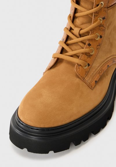 Tan suede ankle boot with black rubber sole, featuring gold eyelets and matching laces. Subtle logo pattern on the side.