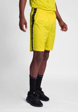 Heldergele sportshorts met zwart chevronpatroon aan de zijkant, elastische tailleband en gladde textuur. Draagt met zwarte sokken en sneakers.