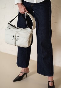 Borsa a spalla grigia chiara con due tasche anteriori, spallacci regolabili e dettagli neri a contrasto, abbinata a jeans a gamba larga di un blu scuro.