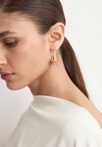 Young woman with long brown hair in a low ponytail wearing shiny gold drop earrings and a white top, facing left against a plain background.