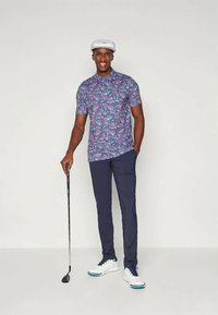 Muž v kvetinovej košeli s krátkym rukávom, tmavomodrých nohaviciach, bielych golfových topánkach a čiapke, usmievajúci sa a drží golfovú palicu, stojí proti jednolitému pozadiu.