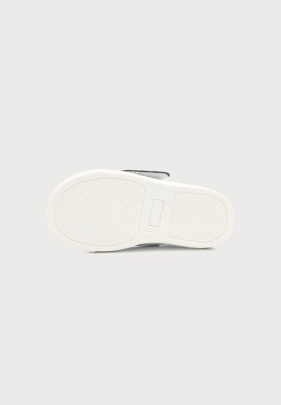 Semelle d’une chaussure en caoutchouc blanche avec un motif de bande de roulement en zigzag, vue de dessous sur un fond gris clair uni.