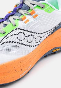 Κοντινό πλάνο ενός λευκού αθλητικού παπουτσιού με πράσινες και πορτοκαλί λεπτομέρειες, γκρι κορδόνια, μαύρο λογότυπο με σχέδιο και πορτοκαλί σόλα με κηλίδες, με την επιγραφή "Vibram".