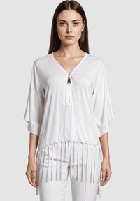 Mujer vestida con una blusa blanca de corte holgado y escote en V, con detalle de cremallera y dobladillo con flecos, combinada con pantalones blancos a juego, de pie frente a un fondo liso.