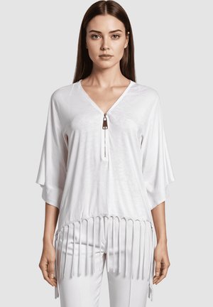 Mujer vestida con una blusa blanca de corte holgado y escote en V, con detalle de cremallera y dobladillo con flecos, combinada con pantalones blancos a juego, de pie frente a un fondo liso.