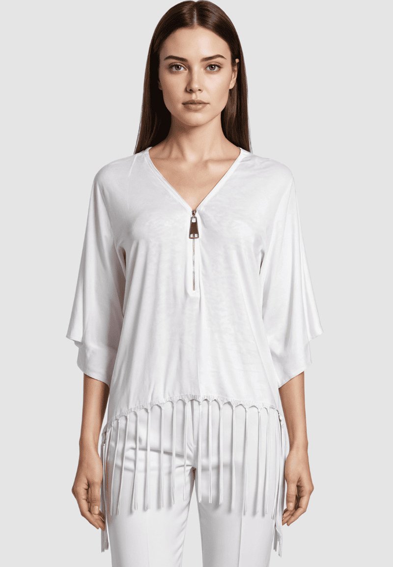 Mujer vestida con una blusa blanca de corte holgado y escote en V, con detalle de cremallera y dobladillo con flecos, combinada con pantalones blancos a juego, de pie frente a un fondo liso.
