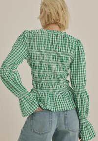 Grøn og hvid gingham-bluse med samlet bodice og lange ærmer. Peplum-kanten og de flæset ærmer tilføjer tekstur og form.