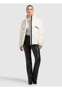 Biała pikowana kurtka z czarnymi akcentami, z zamkiem błyskawicznym z przodu, wysokim kołnierzem i poziomym pikowaniem, noszona z czarnymi skórzanymi spodniami.