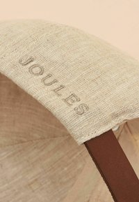 Beigenvärinen kangaslakki pellavapintaisella tekstuurilla ja vaaleanruskealla keinonahkareunuksella. "JOULS" on hienovaraisesti kirjailtu lippaosaan.