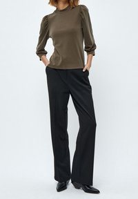 Olivgrön långärmad topp med puffade axlar, kombinerad med svarta vida byxor och svarta spetsiga skor, framför en ljus bakgrund.