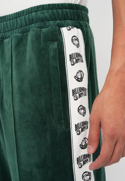 Zaļie velūra sporta bikses ar elastīgu jostu, sānu kabatām un baltu svītru, kurā redzams "Billionaire Boys Club" logo un astronautu grafikas.