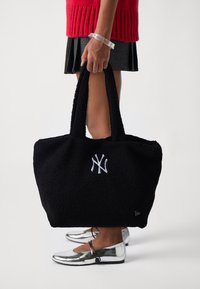 Μαύρη τσάντα tote με υφή από αφράτο ύφασμα, διαθέτοντας λευκό κεντημένο λογότυπο. Δύο ανθεκτικοί ιμάντες, σχεδιασμένοι για μεταφορά.