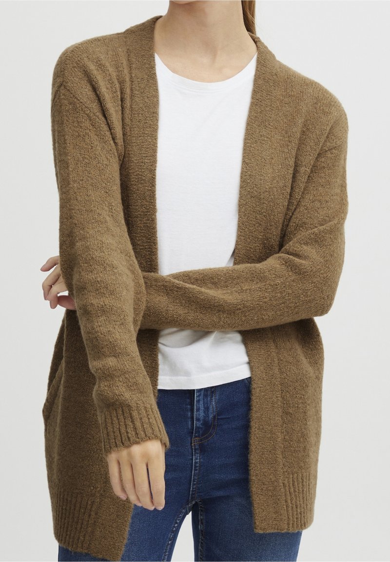 Personne portant un cardigan en tricot marron sur un t-shirt blanc avec un jean bleu, les bras croisés à la taille devant un fond uni.