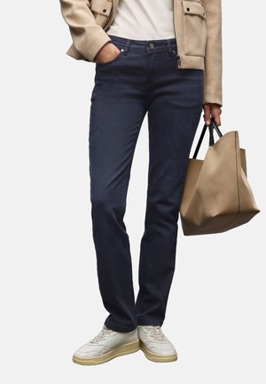 Persoon in donkerblauwe jeans, beige jas, witte sneakers, die een grote beige handtas met zwarte handvatten naast zich houdt.