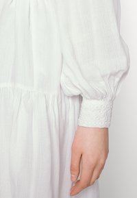 Λευκό φόρεμα με υφασμάτινη υφή, μα gathered μέση και φουσκωτά μανίκια. Οι μανσέτες παρουσιάζουν μια λεπτομερή, κεντητή σχέδια.