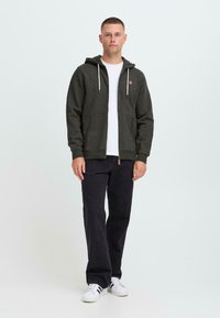 Young man standing wearing dark gray zip-up hoodie, white t-shirt, black pants, and white sneakers with black stripes on white background.