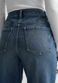 Korkeavyötäröiset siniset farkut, joissa on takataskut, puettuna valkoisen ribbauspaidat pitkällä hihalla, paljastaen alaselän ja vyötärön alueen.