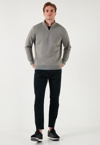 Maglione zip-up grigio chiaro con colletto stand, abbinato a jeans slim-fit neri e sneakers nere. Tessuto liscio, design casual.