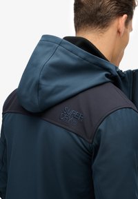 Marineblaue Jacke mit einer strukturierten Kapuze, schwarzen Schultereinsätzen und blauer "SUPERDRY"-Markenkennzeichnung. Der Stoff wirkt glatt und strapazierfähig.