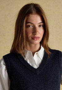 Granatowy wełniany sweter bez rękawów z dekoltem w serek, noszony na białej koszuli z kołnierzykiem. Gładka tekstura z prążkowanymi detalami wzdłuż dekoltu.