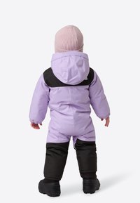 Fialová dětská kombinéza na sníh s černou horní částí, přiléhavou kapucí a elastickými manžetami. Doplněná světle růžovou pletenou čepicí a černými botami.