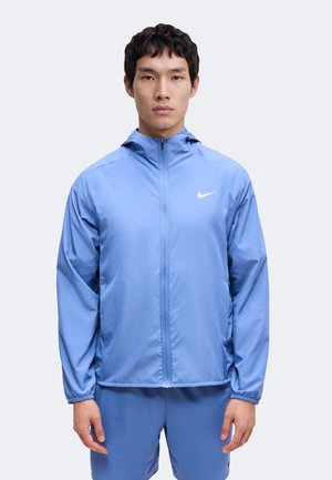 Mladý muž v svetlomodrej Nike bunde na zips s kapucňou a zodpovedajúcich modrých šortkách stojí na obyčajnom pozadí.