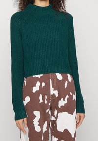Vrouw in een donkergroene geribde trui en een broek in het bruin met witte abstracte vlekken, staande tegen een effen achtergrond.