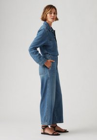 Combinaison en denim bleu moyen avec un haut en veste courte, un pantalon large, une poche poitrine et des coutures contrastées. Portée avec des sandales plates noires.