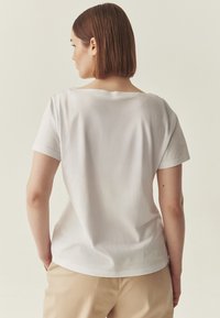 Chemise blanche à manches courtes avec un large décolleté, texture douce, coupe décontractée, couture arrière visible, portée avec un pantalon beige.