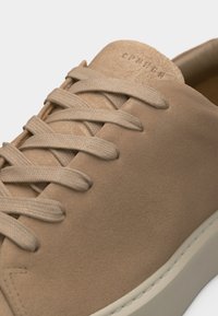 Gros plan sur une basket beige en daim à lacets ton sur ton avec des coutures visibles sur le bord de la semelle.