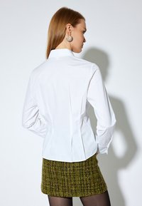Blusa blanca de manga larga con cuello, con pliegues en la parte posterior. Combinada con una minifalda texturizada de patrón verde y marrón.