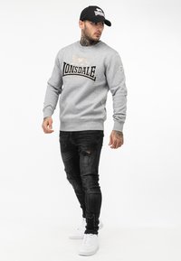Grijze sweatshirt met zwarte "Lonsdale" tekst en leeuwenembleem, gecombineerd met zwarte versleten jeans en witte sneakers; model draagt een zwarte pet.