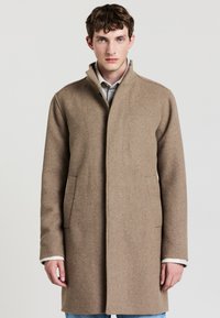Cappotto in lana marrone con colletto alto, dotato di tasche laterali e design pulito e minimalista. Texture morbida e vestibilità su misura.