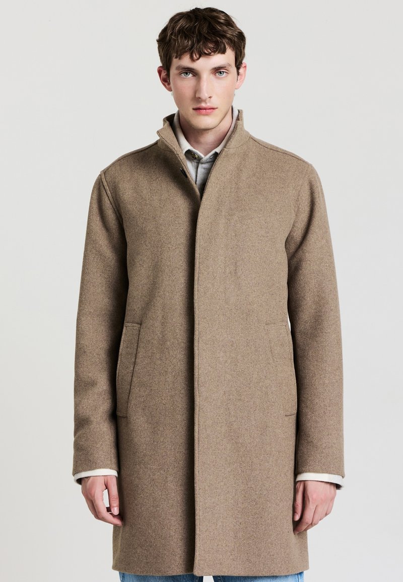 Cappotto in lana marrone con colletto alto, dotato di tasche laterali e design pulito e minimalista. Texture morbida e vestibilità su misura.