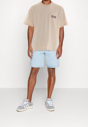 Mann steht und trägt ein beiges Kurzarm-T-Shirt mit dem Schriftzug "PEQUS", hellblaue Jeansshorts und graue und weiße Turnschuhe vor weißem Hintergrund.