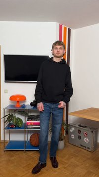 Νέος άνδρας με μαύρο φούτερ με κουκούλα και τζιν που στέκεται σε δωμάτιο με τηλεόραση τοποθετημένη στον τοίχο, πορτοκαλί λάμπα, ραφιέρα με βιβλία, μπάλα του μπάσκετ και βαλίτσα με αυτοκόλλητα.