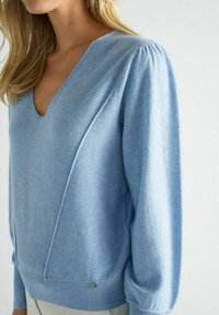Pull en maille bleu clair avec un col en V, des manches bouffantes et un ourlet côtelé. Présente des détails de couture subtils et un petit accent métallique.