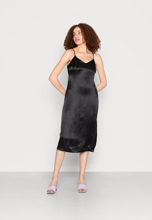 Vestido de seda preto com alças finas; comprimento até a meia panturrilha e textura suave. Combinado com sapatos desportivos lilás num fundo neutro.