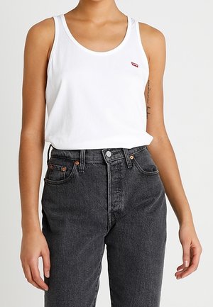 Person iført en hvid ærmeløs tanktop med et lille rødt logo og højtaljede sorte jeans, stående foran en ensfarvet baggrund.