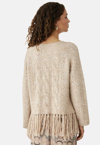 Suéter de punto color beige claro con un patrón de trenzas, que presenta un detalle de flecos cortos en el dobladillo y un corte relajado. Textura suave.