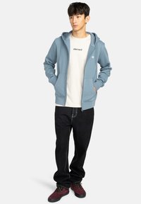 Felpa con zip di colore azzurro chiaro, con cappuccio foderato, due tasche anteriori e un logo circolare sul petto, abbinata a una t-shirt bianca e jeans scuri.