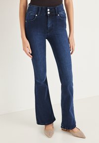 Vaqueros acampanados de mezclilla azul oscuro de talle alto con dos bolsillos delanteros, cierre de botones y un sutil desgaste en los muslos. Se llevan zapatos planos.