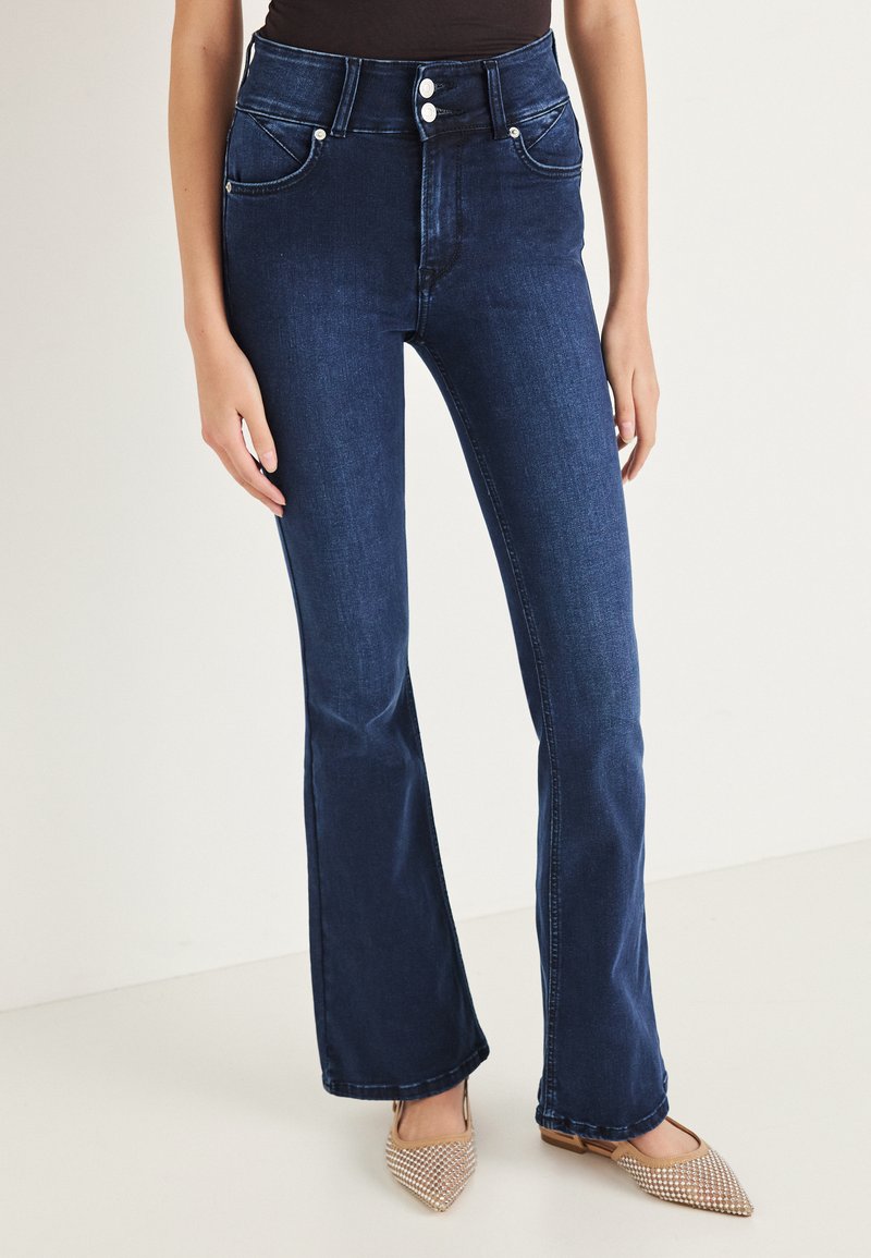 Vaqueros acampanados de mezclilla azul oscuro de talle alto con dos bolsillos delanteros, cierre de botones y un sutil desgaste en los muslos. Se llevan zapatos planos.