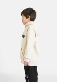 Felpa beige con tasca frontale e scritte nere, indossata su pantaloni neri con cerniere. La felpa presenta un grande cappuccio e polsini a costine.